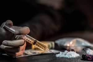 Male junkie hand holding drug injection syringe while lying near heroin powder, spoon and cigarette lighter for heroin cooking and money on dark floor. Hard drug addiction concept